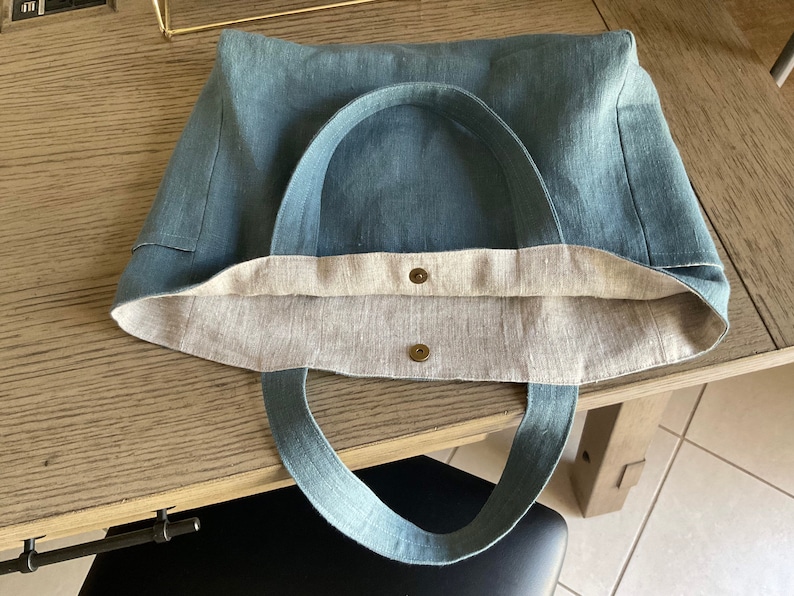 May include: A blue linen tote bag with two brown metal snaps and a light brown linen lining. The bag has two long blue linen straps.