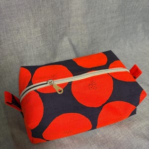 May include: A rectangular zippered pouch with a vibrant pattern of large red circles on a navy blue background. The pouch has a silver zipper and small red fabric loops on each end. The overall design is eye-catching and modern.