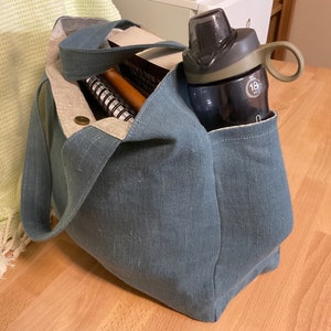 May include: A blue canvas tote bag with a water bottle in the side pocket. The bag is filled with books and a notebook.