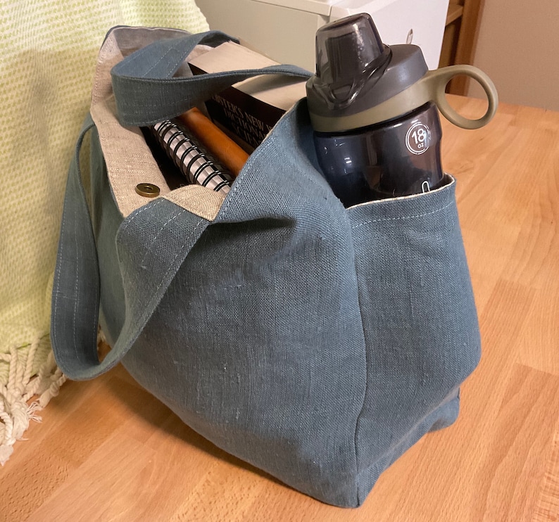 May include: A blue canvas tote bag with a water bottle in the side pocket. The bag is filled with books and a notebook.