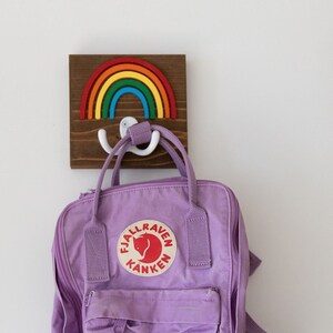 Pode incluir: Uma mochila Fj&auml;llr&auml;ven K&aring;nken roxa pendurada em um gancho de madeira em forma de arco-&iacute;ris. O gancho tem um gancho duplo de pl&aacute;stico branco.