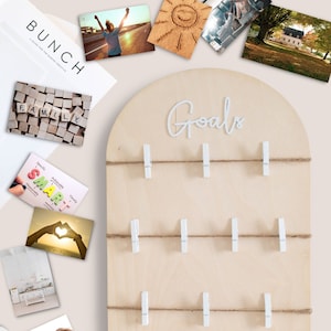 May include: A light wood goal board with the word "Goals" written in white letters. The board has 10 white clothespins attached to twine strung across the board. There are several images and papers on a white surface around the board, including a notebook with the word "manifest" written on it.