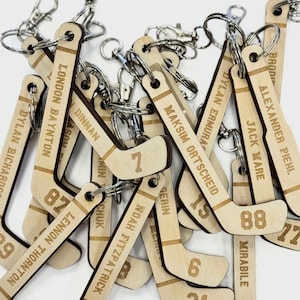 May include: A selection of wooden hockey stick keyrings. Each keyring is engraved with a name and a number. The keyrings are made of light-coloured wood and have silver key rings attached.