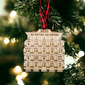May include: A wooden Christmas ornament shaped like a hockey team jersey with the text "US Okotoks Big Rippers" and the year "20:23". The ornament features the names and jersey numbers of 11 players.