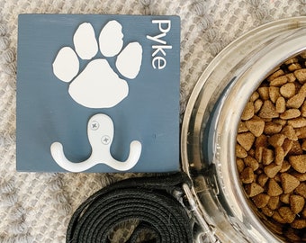 Puede incluir: Un soporte de correa para perros de madera azul con una huella de pata blanca y el nombre "Pyke". Un gancho blanco está debajo de la huella de la pata. Una correa negra y un cuenco plateado para perros lleno de pienso también están en la imagen.
