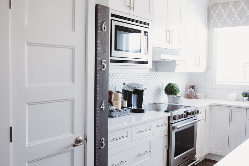 Puede incluir: Una cocina blanca con una tabla de crecimiento en blanco y negro en la pared, un microondas, una cafetera y un horno blanco. La cocina tiene gabinetes blancos y una encimera blanca.