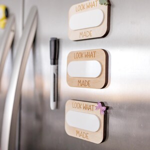 May include: Three wooden signs with white dry erase surfaces and the text "Look What I Made" on each sign. The signs are attached to a stainless steel refrigerator with magnets. The magnets are a green dinosaur, a white flower, and a pink bow.