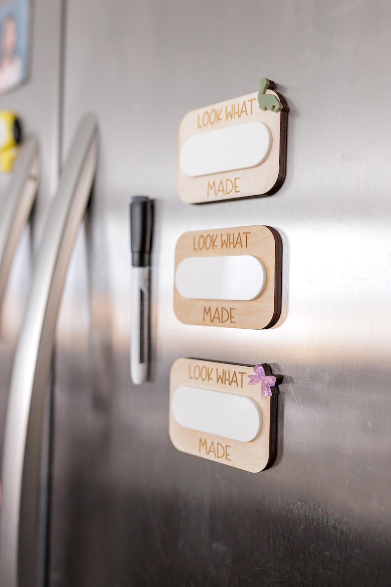 May include: Three wooden signs with white dry erase surfaces and the text "Look What I Made" on each sign. The signs are attached to a stainless steel refrigerator with magnets. The magnets are a green dinosaur, a white flower, and a pink bow.