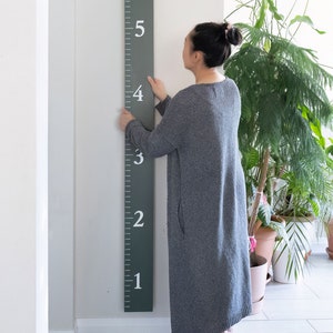 May include: A person stands next to a green wooden growth chart with white numbers 1 through 6. The chart is mounted on a white wall.