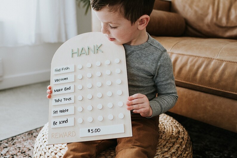 May include: A wooden chore chart with the name "HANK" at the top. The chart has a list of chores, including "Feed Fish", "Vacuum", "Make Bed", "Tidy Toys", "Set table", and "Be kind". There are white circles next to each chore, and a reward of $5.00 is listed at the bottom.