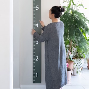 Puede incluir: Una persona se encuentra junto a una tabla de crecimiento de madera verde con n&uacute;meros blancos del 1 al 6. La tabla est&aacute; montada en una pared blanca.