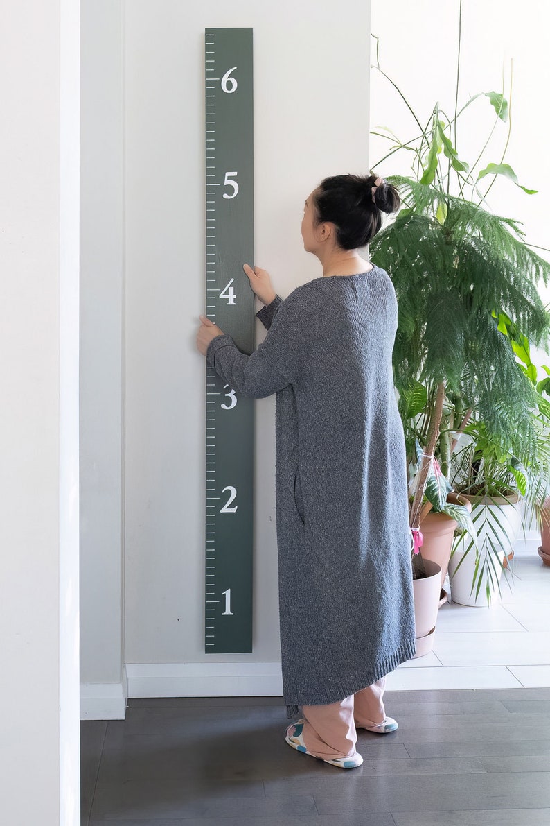 Puede incluir: Una persona se encuentra junto a una tabla de crecimiento de madera verde con n&uacute;meros blancos del 1 al 6. La tabla est&aacute; montada en una pared blanca.