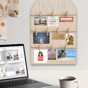May include: A wooden wall decor with a white arch and the text "Manifest it" at the top. The arch has clips holding various cards with inspirational messages and images, including "Thankful Grateful Blessed", "Faith", "Focus", "Just Start", and a picture of a tropical beach.