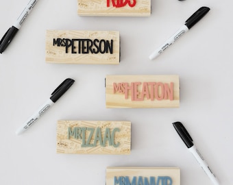 May include: Five rectangular wooden stamps with raised lettering in red, black, pink, green, and blue. The stamps feature names including TOPP KIDS, MRS PETERSON, MS HEATON, MR IZAAC, and MR MANVIR, surrounded by black dry erase markers.
