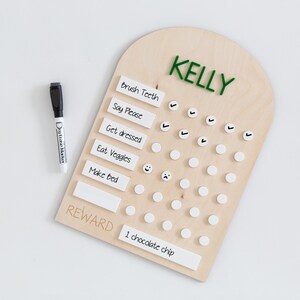 May include: A wooden chart with the name "KELLY" at the top. The chart has a list of chores for children, such as "Brush Teeth", "Say Please", "Get Dressed", "Eat Veggies", and "Make Bed". There are white circles next to each chore, and a check mark can be placed in the circle when the chore is completed. The chart also has a section for "REWARD" with the text "1 chocolate chip".
