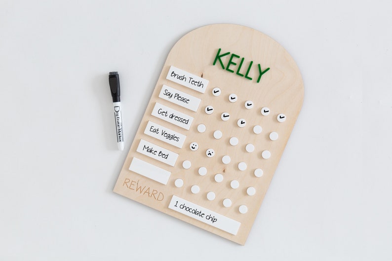May include: A wooden chart with the name "KELLY" at the top. The chart has a list of chores for children, such as "Brush Teeth", "Say Please", "Get Dressed", "Eat Veggies", and "Make Bed". There are white circles next to each chore, and a check mark can be placed in the circle when the chore is completed. The chart also has a section for "REWARD" with the text "1 chocolate chip".