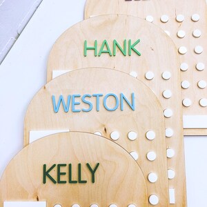 May include: Four wooden arch-shaped boards with white circles and names written in colorful letters. The names are Ella, Hank, Weston, and Kelly.