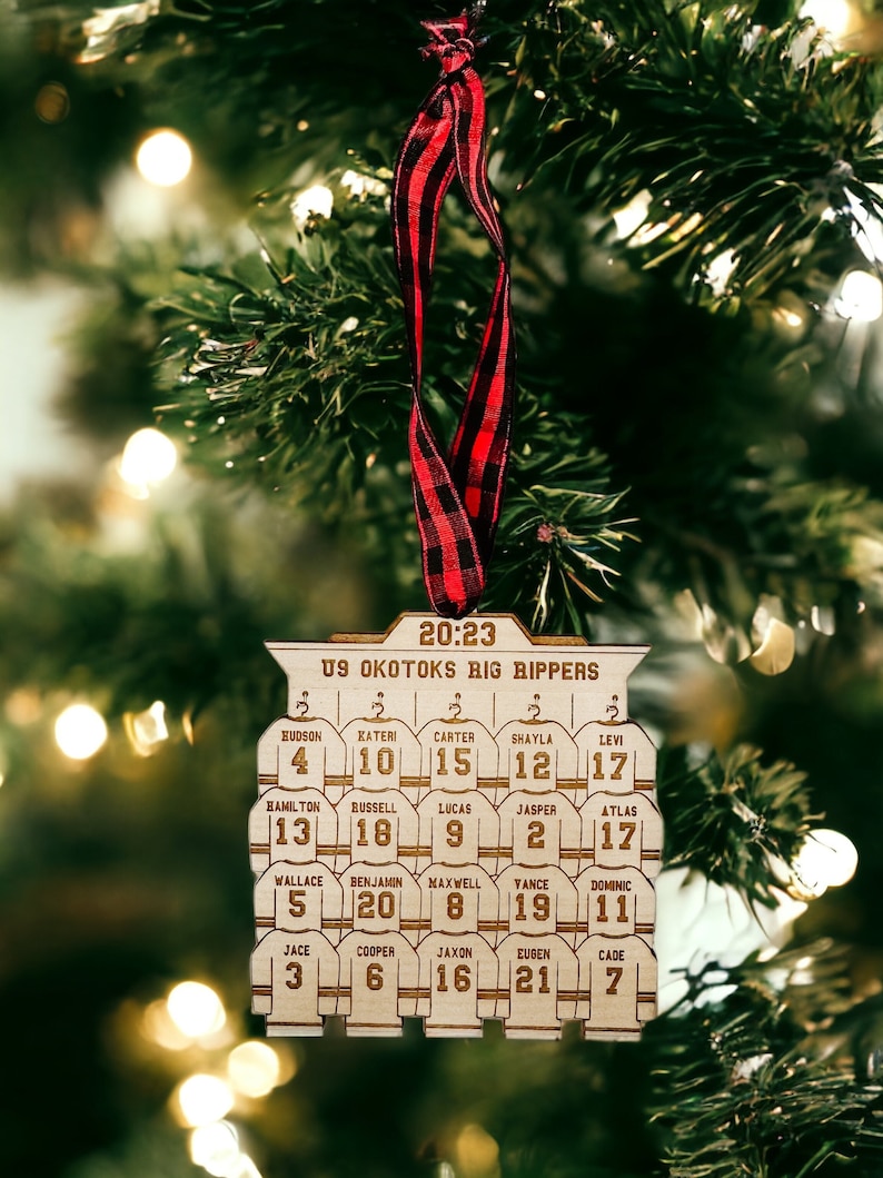 May include: A wooden Christmas ornament shaped like a hockey team jersey with the text "US Okotoks Big Rippers" and the year "20:23". The ornament features the names and jersey numbers of 11 players.