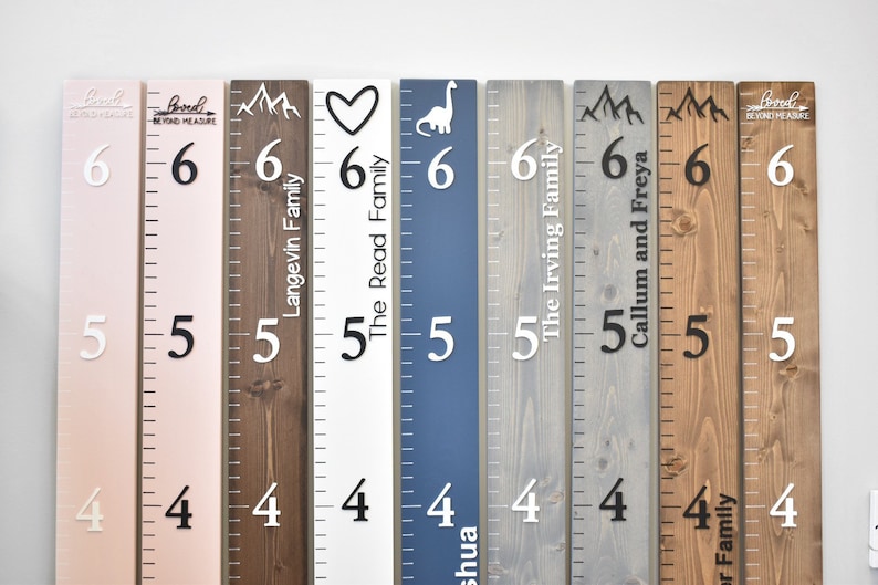 May include: Eight wooden growth charts with mountain, heart, dinosaur, and arrow designs. The charts are painted in various colors, including pink, brown, white, blue, gray, and natural wood. Each chart has a name and family name printed on it, such as "Langevin Family" and "The Irving Family".