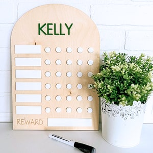 May include: A wooden chart with a rounded top, labeled "KELLY" in green letters. The chart has white dry-erase spaces for writing tasks and a reward section. There are 35 white circles for marking off completed tasks.