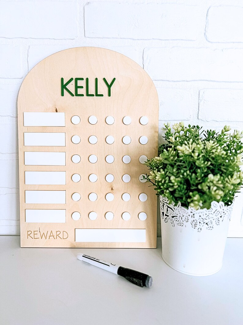 May include: A wooden chart with a rounded top, labeled "KELLY" in green letters. The chart has white dry-erase spaces for writing tasks and a reward section. There are 35 white circles for marking off completed tasks.