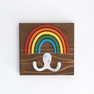 Pode incluir: Um gancho de parede de madeira em forma de arco-&iacute;ris com um gancho duplo branco. O arco-&iacute;ris &eacute; feito de madeira pintada de vermelho, laranja, amarelo, verde, azul e roxo.