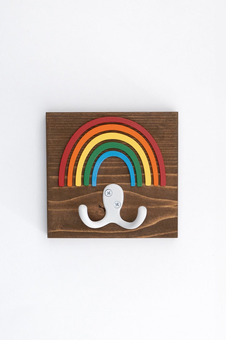 Pode incluir: Um gancho de parede de madeira em forma de arco-&iacute;ris com um gancho duplo branco. O arco-&iacute;ris &eacute; feito de madeira pintada de vermelho, laranja, amarelo, verde, azul e roxo.