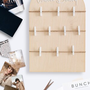 May include: A wooden vision board with the words "Vision Board" written in white script. The board has a string with white clothespins attached to it. There are several magazines, photos, and other items on the table around the board.