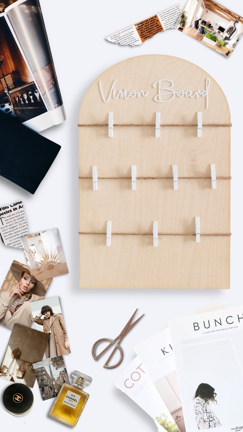 May include: A wooden vision board with the words "Vision Board" written in white script. The board has a light brown wood grain and is decorated with white clothes pegs and twine. The board is surrounded by various items, including magazines, photos, and a perfume bottle.