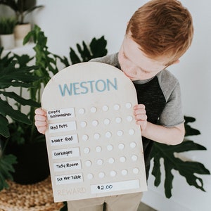 May include: A wooden chore chart with the name Weston at the top. The chart has a list of chores with white circles next to each one. A young boy is holding the chart and looking at it. The chart has a reward section that says $2.00.