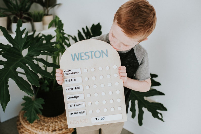 May include: A wooden chore chart with the name Weston at the top. The chart has a list of chores with white circles next to each one. A young boy is holding the chart and looking at it. The chart has a reward section that says $2.00.