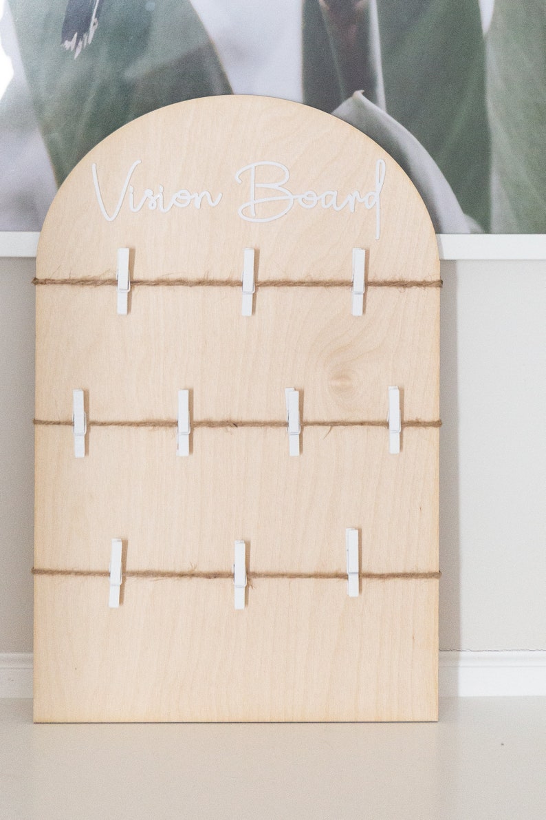 May include: A light wood vision board with the words "Vision Board" written in white lettering. The board has multiple rows of twine with white clothespins attached.