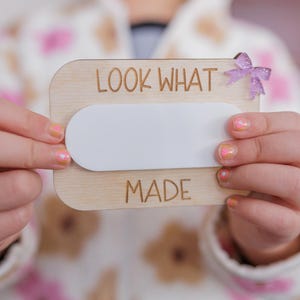 May include: A wooden sign with the text "LOOK WHAT I MADE" and a blank white space in the middle. A child is holding the sign in front of them.