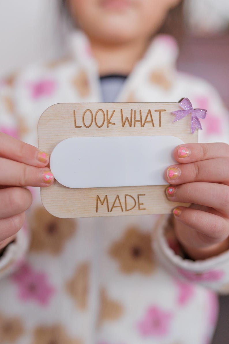 May include: A wooden sign with the text "LOOK WHAT I MADE" and a blank white space in the middle. A child is holding the sign in front of them.