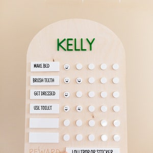 May include: A wooden chart with the name "KELLY" at the top. The chart has a list of chores with smiley faces and blank circles for marking completion. The chores are "Make Bed", "Brush Teeth", "Get Dressed", and "Use Toilet". The chart also has a section for "Reward" with the text "Lollipop or Sticker".