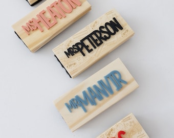 May include: Five rectangular wooden stamps with black felt bases. Each stamp features a light wood handle engraved with school-themed icons like buses and rulers. Raised, colored plastic lettering spells out MR ISAAC, MS HEATON, MRS PETERSON, MR MANVIR, and TOPP KIDS.