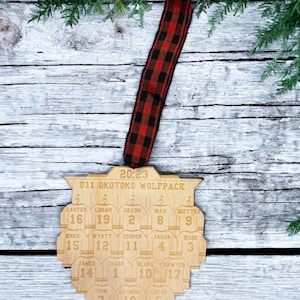 May include: A wooden hockey team ornament with the text "20:23 U11 OKOTOKS WOLFPACK" and the names and numbers of the players on the team. The ornament is hanging from a red and black buffalo plaid ribbon.