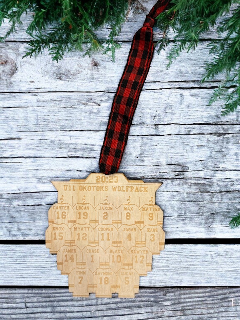 May include: A wooden hockey team ornament with the text "20:23 U11 OKOTOKS WOLFPACK" and the names and numbers of the players on the team. The ornament is hanging from a red and black buffalo plaid ribbon.