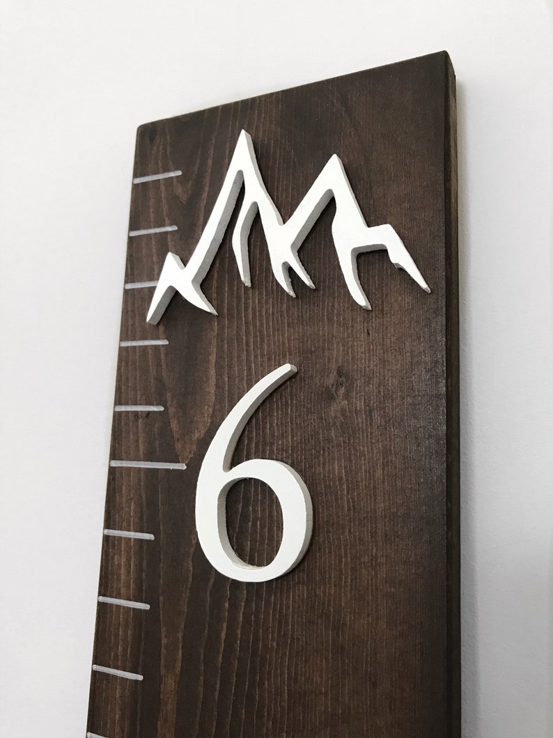 Puede incluir: Un gr&aacute;fico de crecimiento de madera marr&oacute;n oscuro con picos de monta&ntilde;as pintados de blanco y un n&uacute;mero "6" blanco en la parte inferior. El gr&aacute;fico tiene l&iacute;neas blancas para medir la altura.