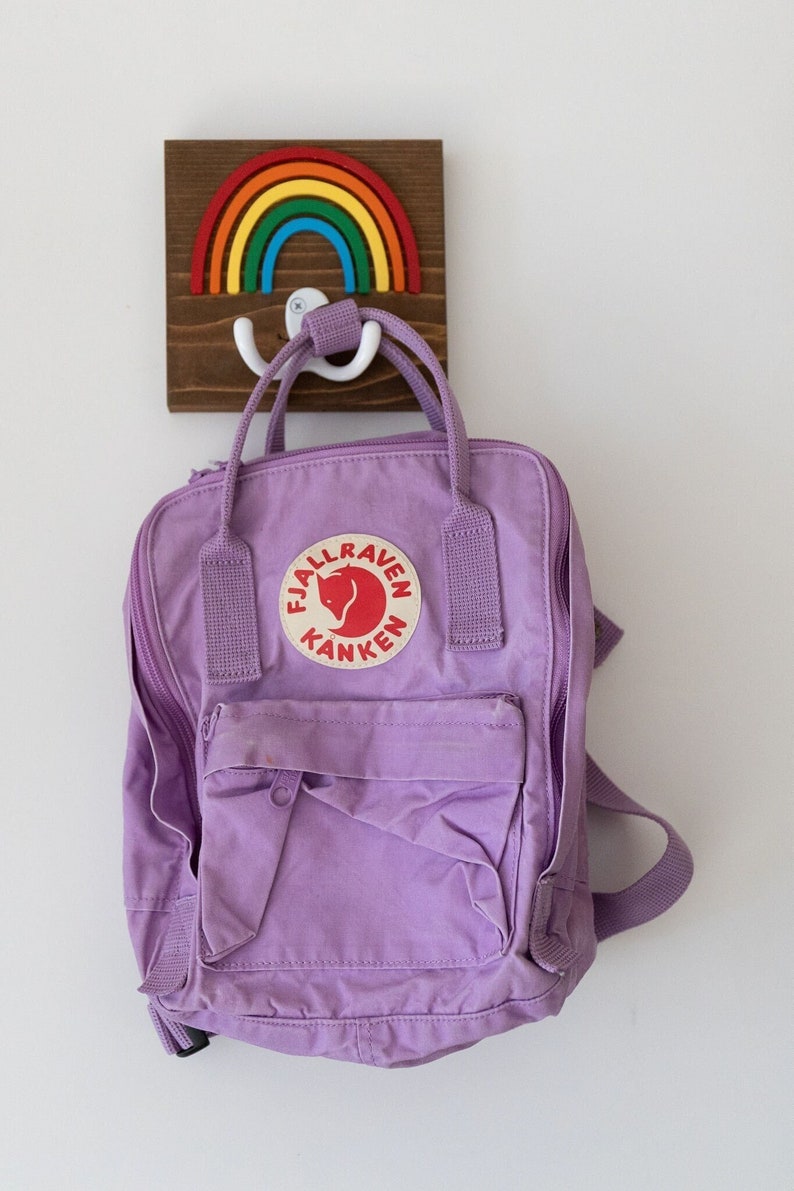 Pode incluir: Uma mochila Fj&auml;llr&auml;ven K&aring;nken roxa pendurada em um gancho de madeira em forma de arco-&iacute;ris. O gancho tem um gancho duplo de pl&aacute;stico branco.