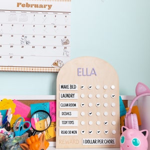 May include: A wooden chore chart with the name "ELLA" at the top. The chart has a list of chores with checkboxes next to each one. The chores are: Make Bed, Laundry, Clean Room, Dishes, Tidy Toys, Read 30 Min. At the bottom of the chart, it says "REWARD I DOLLAR PER CHORE".