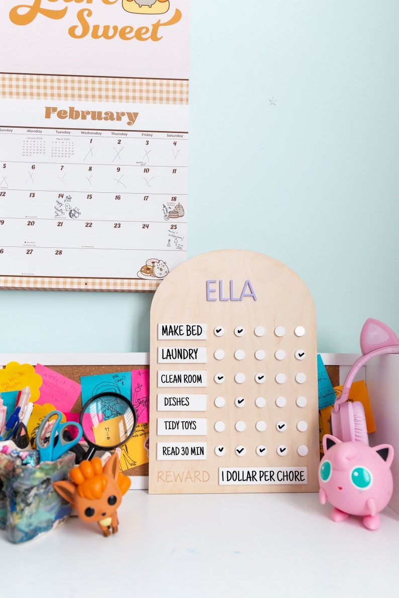 May include: A wooden chore chart with the name "ELLA" at the top. The chart has a list of chores with checkboxes next to each one. The chores are: Make Bed, Laundry, Clean Room, Dishes, Tidy Toys, Read 30 Min. At the bottom of the chart, it says "REWARD I DOLLAR PER CHORE".