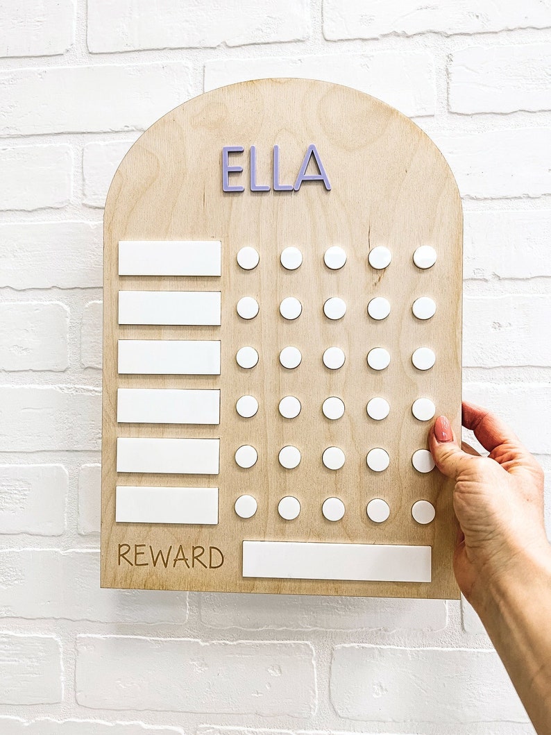 May include: A wooden chart with the name "ELLA" at the top. The chart has five blank white spaces for writing and a grid of white circles for tracking progress. The chart also has a space labeled "REWARD" for writing a reward.