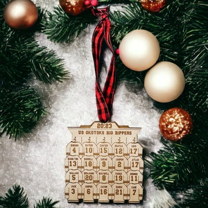 May include: A wooden hockey ornament with the text "20:23 U9 Okotoks Big Rippers" and the names and numbers of a hockey team. The ornament is decorated with a red and black plaid ribbon and is surrounded by gold and brown Christmas ornaments.