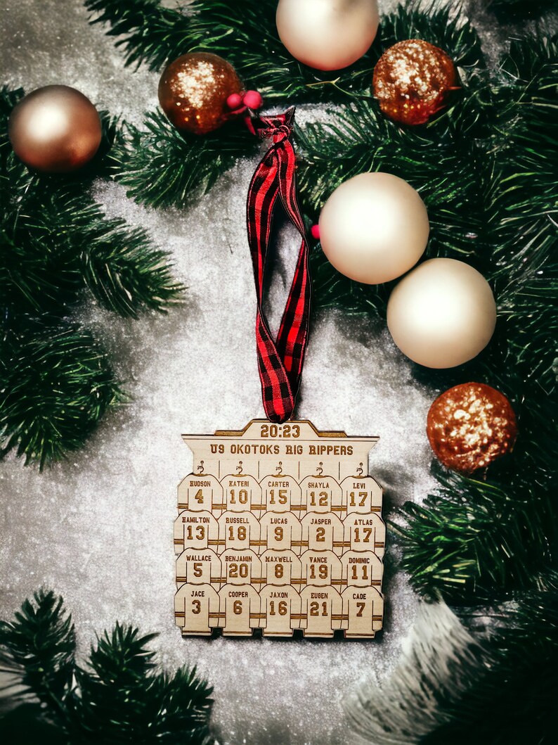 May include: A wooden hockey ornament with the text "20:23 U9 Okotoks Big Rippers" and the names and numbers of a hockey team. The ornament is decorated with a red and black plaid ribbon and is surrounded by gold and brown Christmas ornaments.