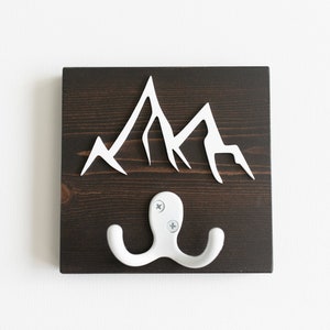 Pode incluir: Um gancho de parede de madeira castanho escuro com uma silhueta de montanha branca e um gancho duplo branco.