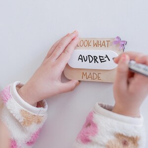 May include: A wooden sign with the text "Look What Audrey Made" and a purple butterfly decoration. The sign is being held by a child's hands.