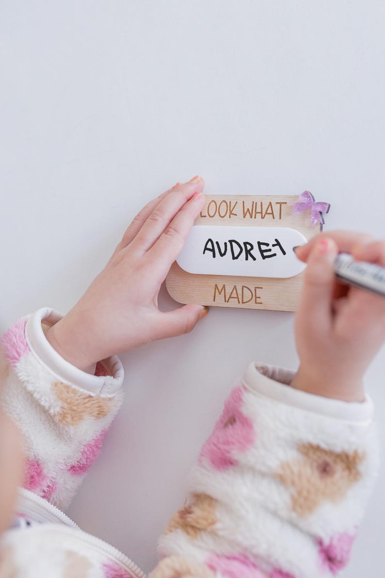 May include: A wooden sign with the text "Look What Audrey Made" and a purple butterfly decoration. The sign is being held by a child's hands.