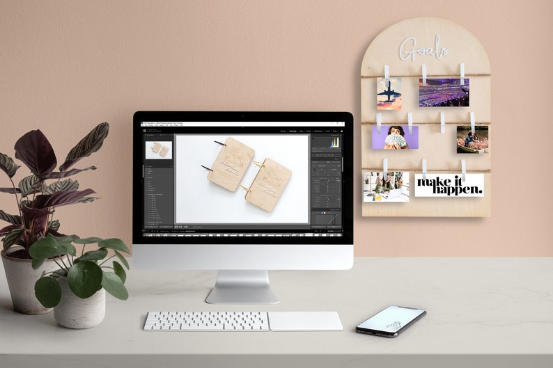 May include: A light wood desktop with a computer monitor displaying a photo of two wooden notebooks. A white keyboard, a black smartphone, and two potted plants are also on the desk. A light wood wall mounted goal board with photos and the text "Goals" and "make it happen." is on the wall behind the desk.