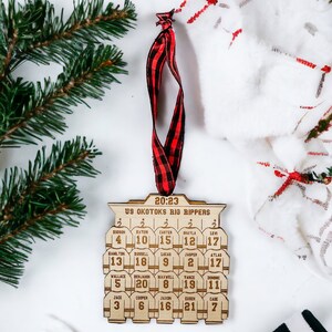 May include: A wooden hockey ornament with the text "20:23 U9 Okotoks Big Rippers" and the names and numbers of a hockey team. The ornament is brown and has a red and black checkered ribbon attached.